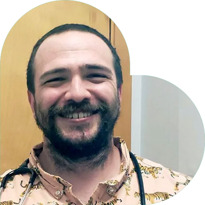 Smiling man with short hair and a beard wearing a tiger-patterned shirt and a stethoscope around his neck, stands in front of a wooden door, exuding the confidence of a dedicated veterinarian.