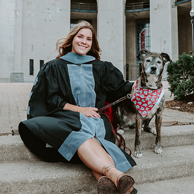 A woman in a graduation gown sits on steps next to a dog wearing a patterned bandana. Both are smiling towards the camera, with a large building in the background.